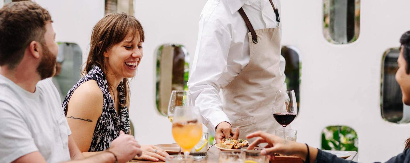 La imagen muestra el entorno de un restaurante. Un camarero está sirviendo a los comensales en la mesa.