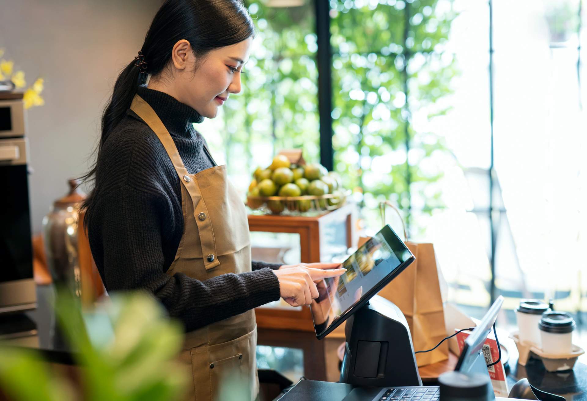 Imagen que muestra a una mujer con un suéter de cuello alto negro y un delantal café usando una tableta montada en un restaurante.