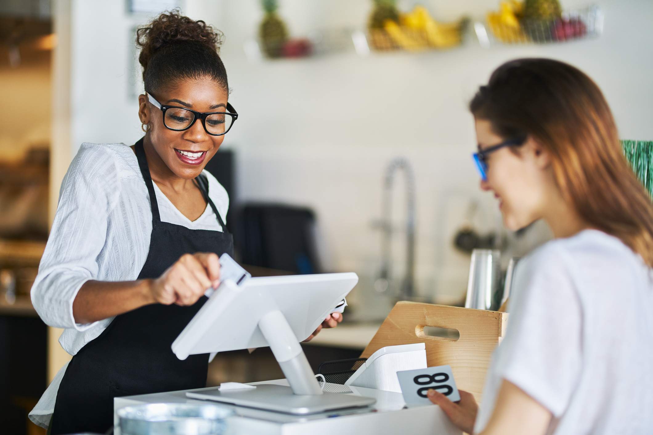 La imagen muestra a una mesera con camisa blanca, delantal negro y lentes con montura negra que pasa la tarjeta de crédito de una clienta en una terminal de pago. La clienta sonríe mientras espera. 