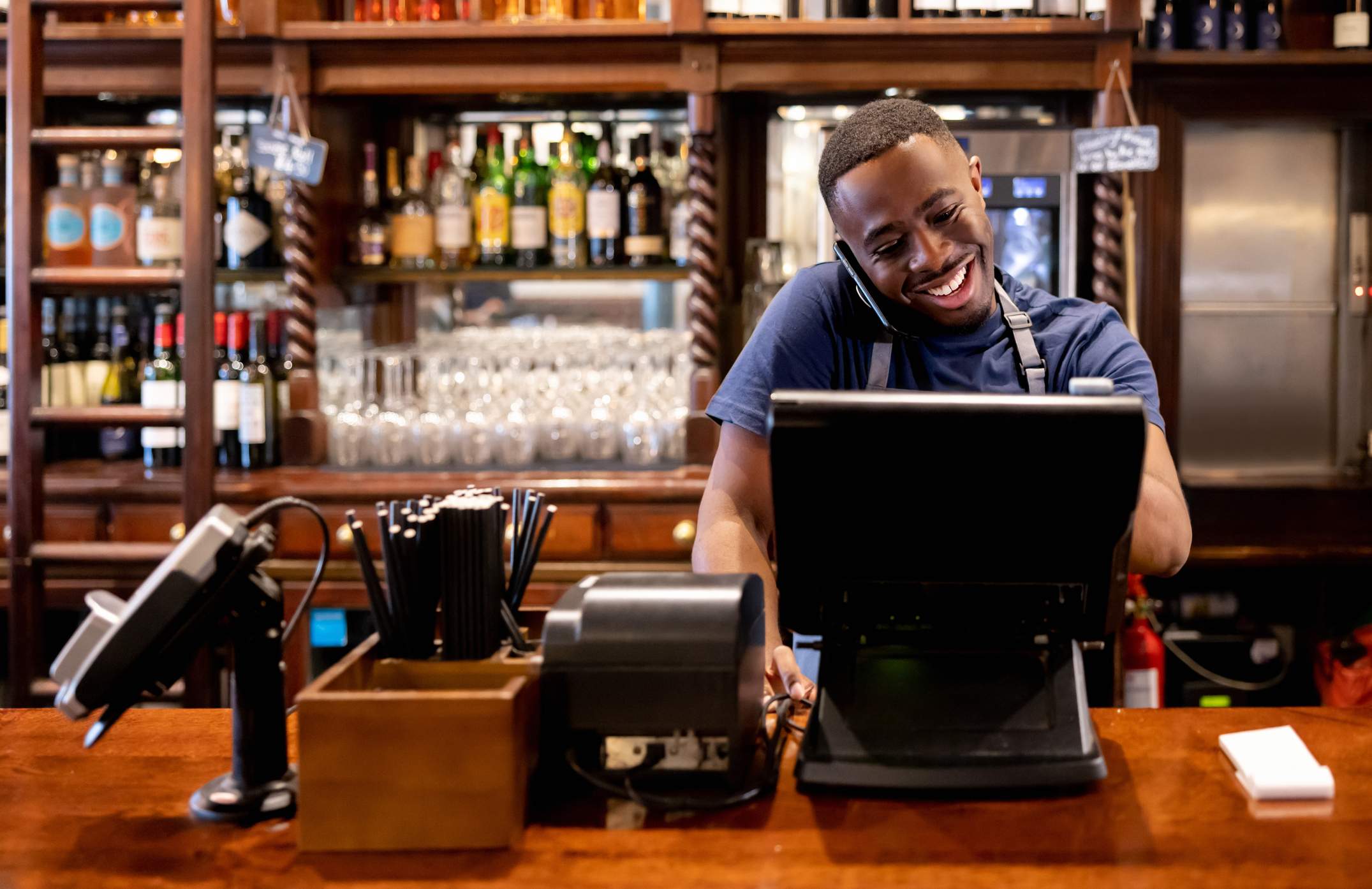Imagen que muestra a un mesero sonriente con camiseta azul y un delantal negro hablando por teléfono mientras usa una computadora situada frente a él. Hay un bar con repisas y botellas en el fondo.