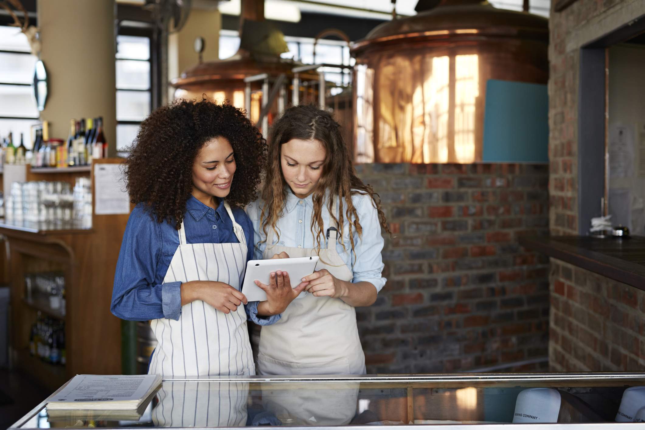 La imagen muestra a dos trabajadores de un restaurante. Una sostiene una tableta y le muestra a la otra mesera algo en la tableta.