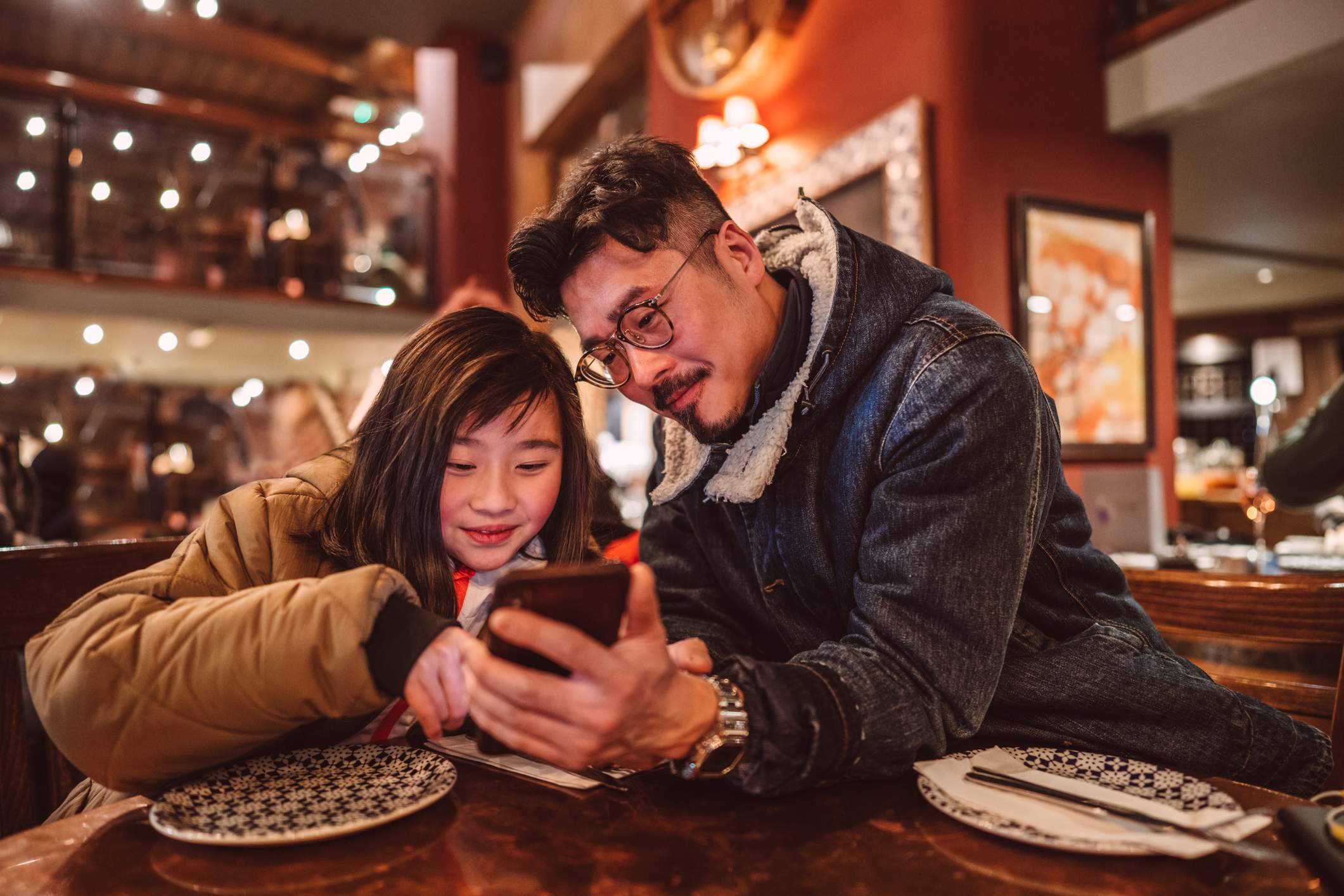 La imagen muestra a una niña y a su padre usando juntos un teléfono mientras están sentados en la mesa de un restaurante. 