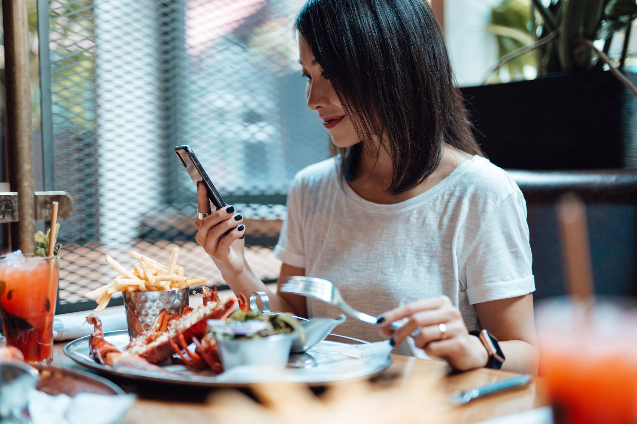 La imagen muestra a una persona sonriendo a su teléfono mientras está sentada en un restaurante Delante de ella hay una bebida de colores, un plato con mariscos y una porción de papas a la francesa. 
