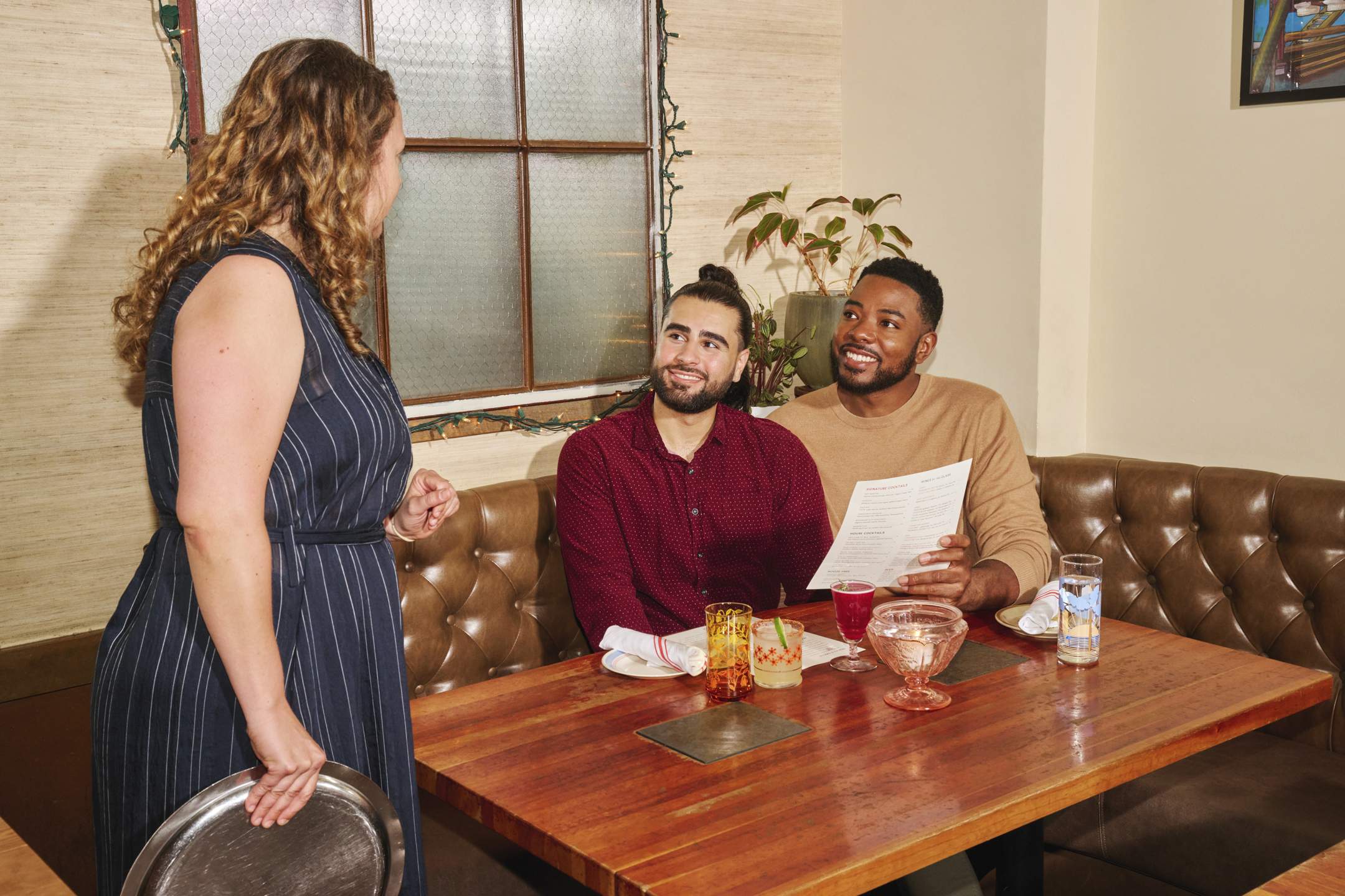 la imagen muestra un entorno de restaurante. Dos comensales están consultando un menú mientras una mesera está de pie junto a la mesa.