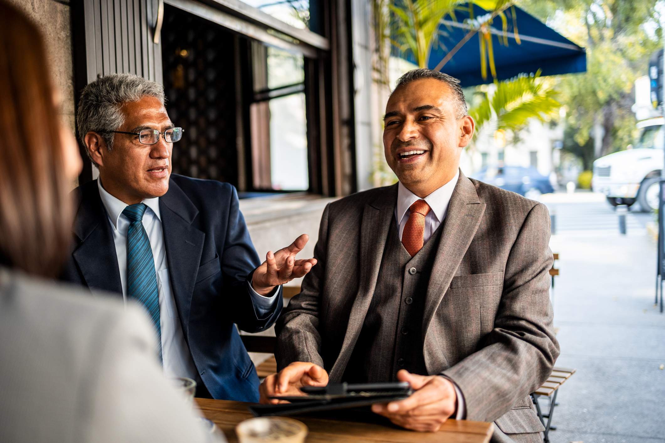 En esta foto, dos hombres de negocios están sentados fuera de un restaurante y hablan con una tercera persona. Llevan ropa de oficina y uno de ellos sostiene una tableta en la mano.