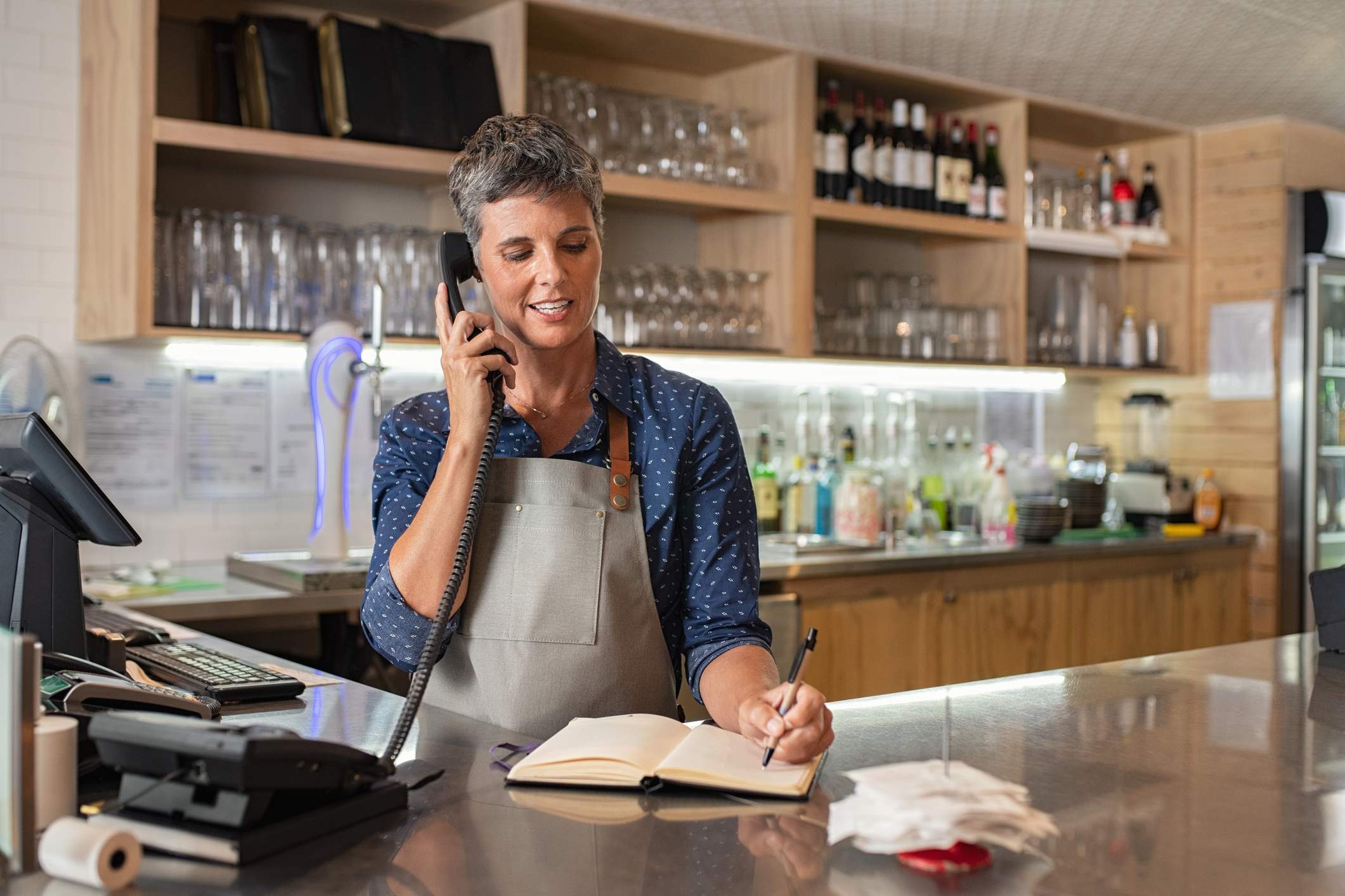La imagen muestra a una trabajadora de restaurante en la recepción de un restaurante, hablando por teléfono mientras toma notas en un bloc de notas.